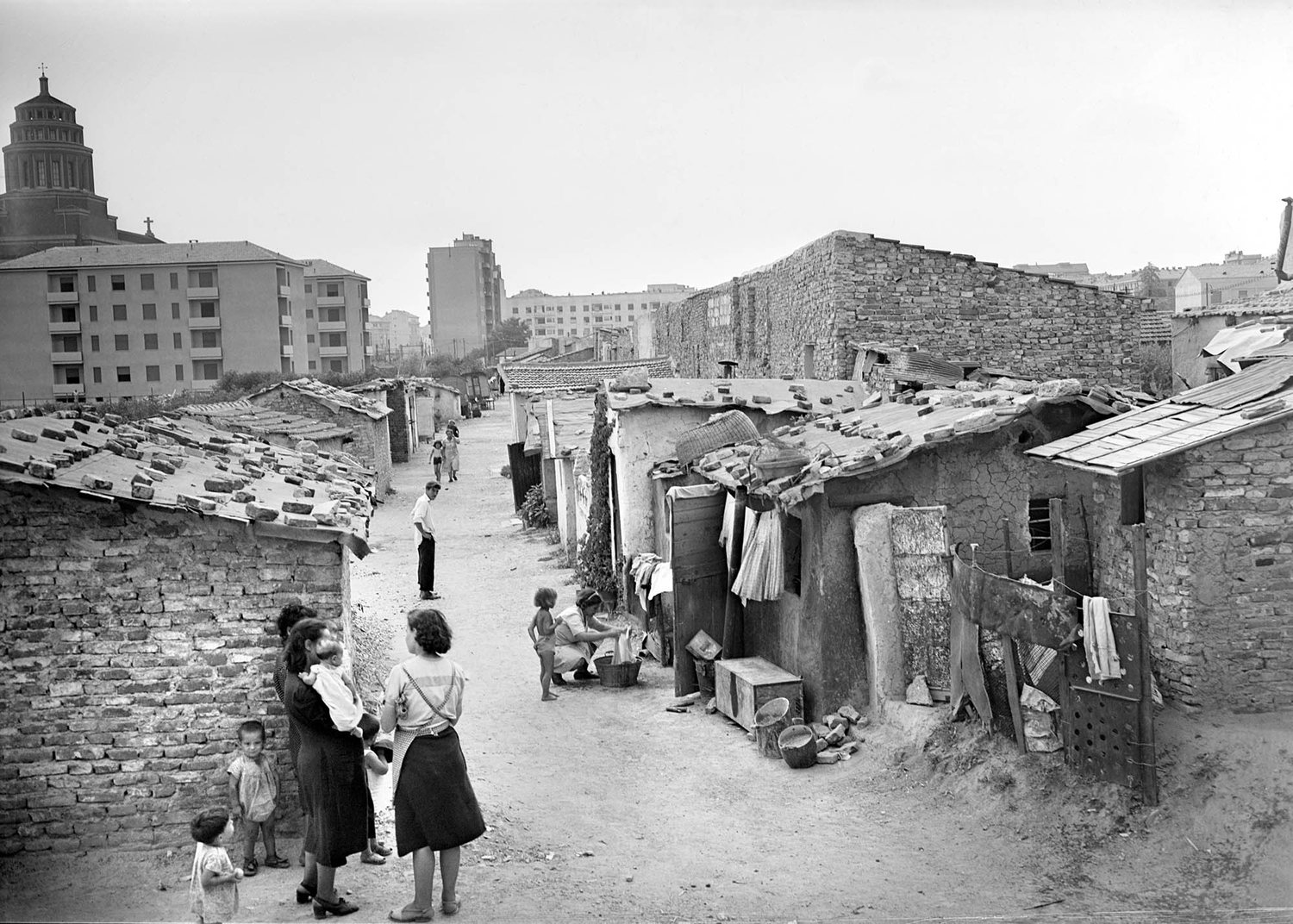 Le baraccopoli di via Marescalchi nel 1950: in primo piano alcune donne e bambini tra le baracche di mattoni lungo una via sterrata; sullo sfondo i grandi caseggiati moderni e, sulla sinistra, una chiesa con la cupola. Fotografia Publifoto.