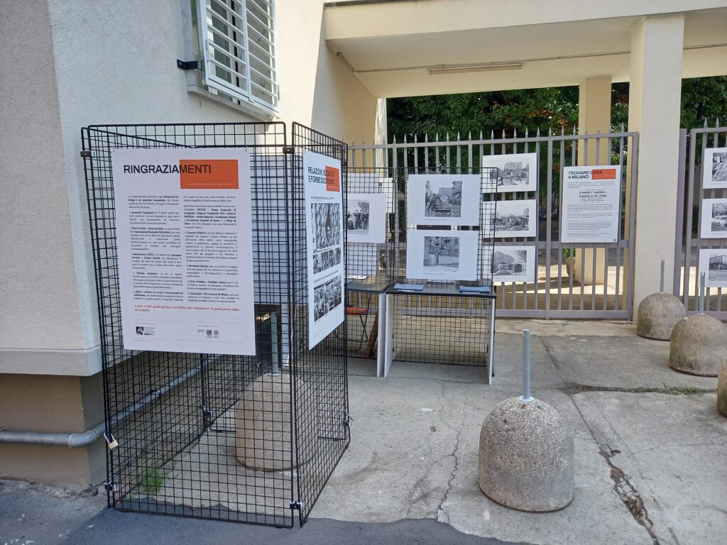 Gli espositori con i pannelli "Ringraziamenti" e "Relazioni, socialità e forme di comunità", con un tavolo e, sullo sfondo, le fotografie appese alla cancellata