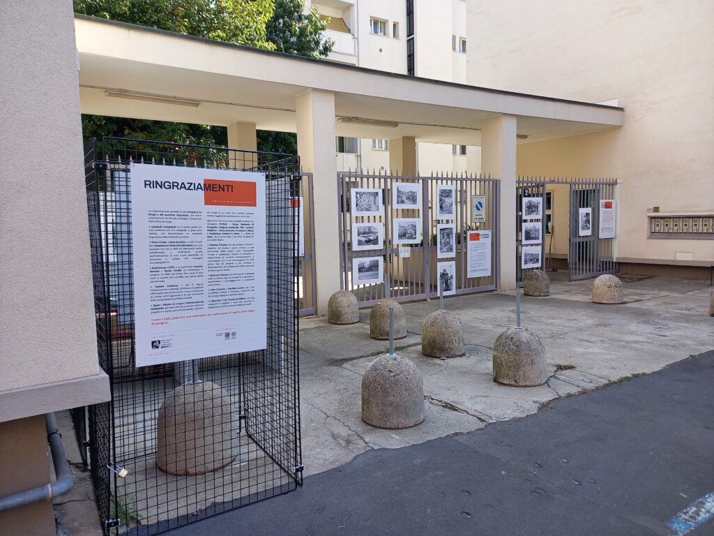 In primo piano il pannello "Ringraziamenti" su un espositore a griglia; dietro, le fotografie della mostra appese alla cancellata d'ingresso