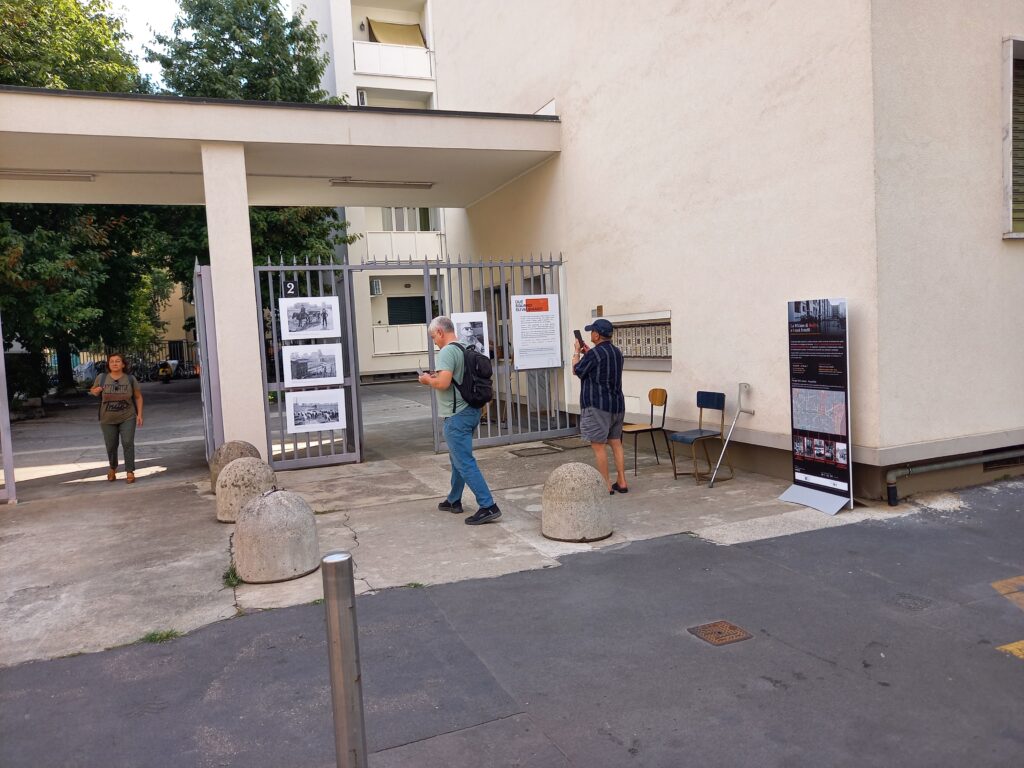 Due uomini vicino all'ingresso: uno legge i pannelli, l'altro li fotografa; sulla destra il totem della mostra.
