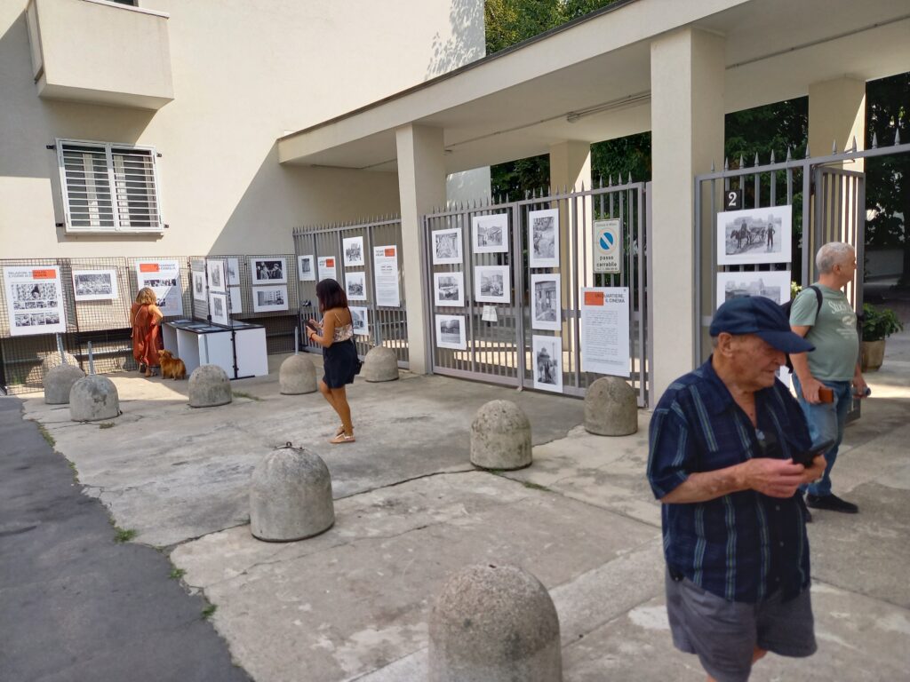 Visitatori davanti ai pannelli sulla cancellata; in primo piano un uomo con il telefono, più indietro una persona con un cane.