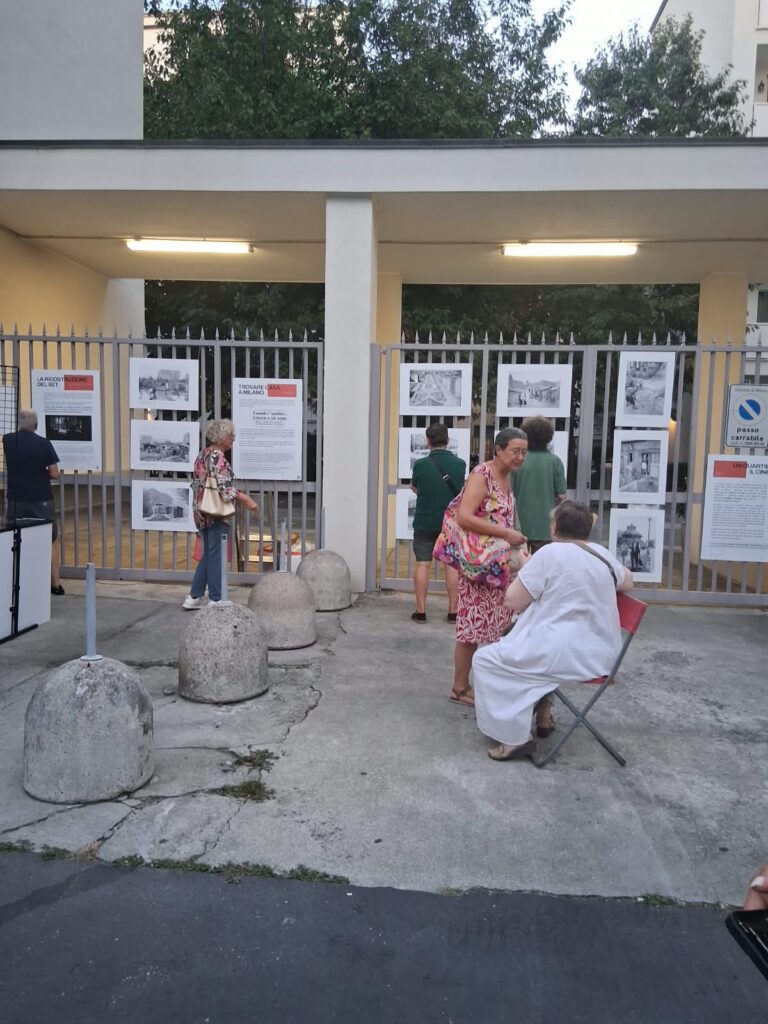 Verso sera, alcune persone davanti alla mostra; una donna è seduta su uno sgabellino pieghevole rosso.