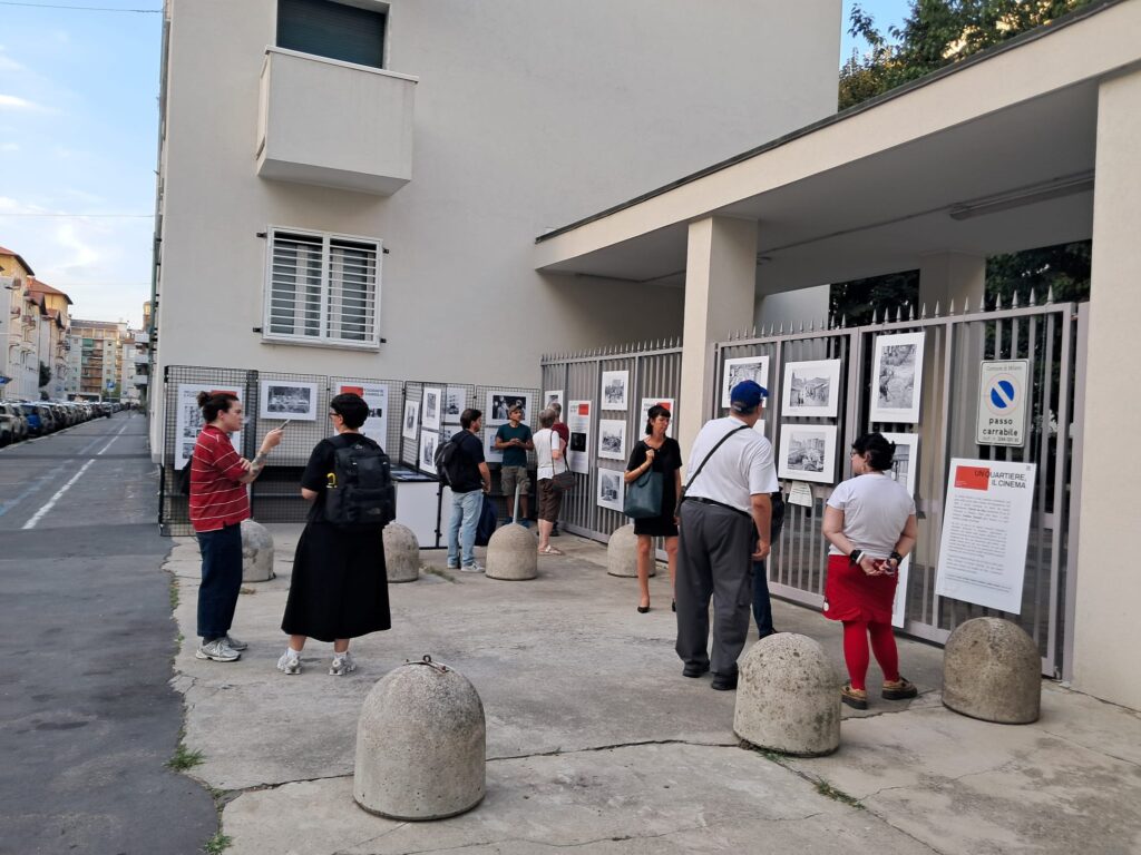 Due persone conversano in primo piano mentre altri visitatori guardano le fotografie lungo la via.